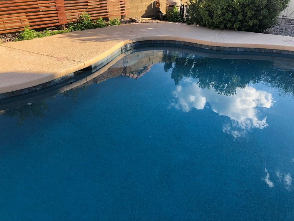 Clear swimming pool reflecting blue sky and clouds, surrounded by greenery and stone patio.