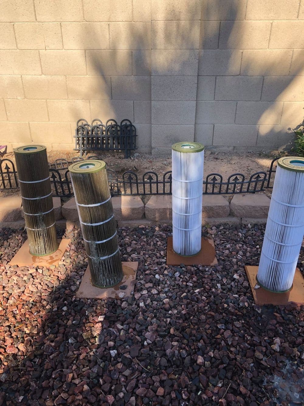 Four cylindrical pool filters standing on a gravel surface against a concrete wall.